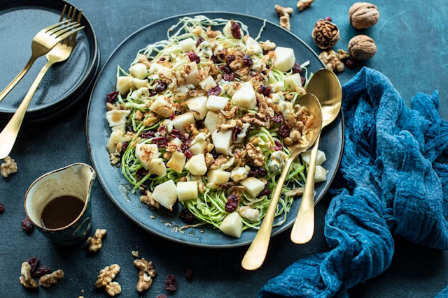 A top-down view of a large teal ceramic platter filled with a shaved Brussels sprout salad, sliced pears, walnuts, dried cranberries, and blue cheese crumbles on a dark blue surface with gold utensils.