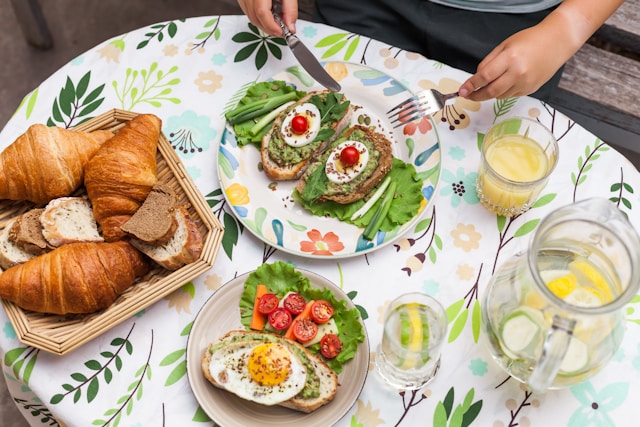 A top-down view of a brunch table with a green floral tablecloth, featuring avocado toast with a boiled egg, a fried egg on sourdough with tomatoes, a basket of croissants, and lemon water.