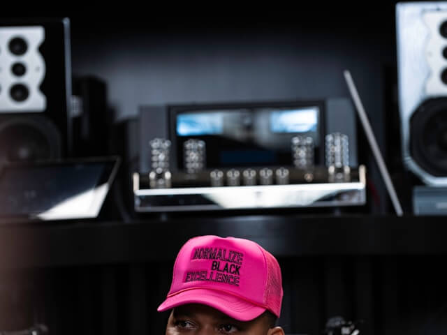 A close-up of a person wearing a vibrant neon pink trucker hat with the phrase "NORMALIZE BLACK EXCELLENCE" embroidered in black block letters. The background is a blurred high-end audio studio with speakers and sound equipment.