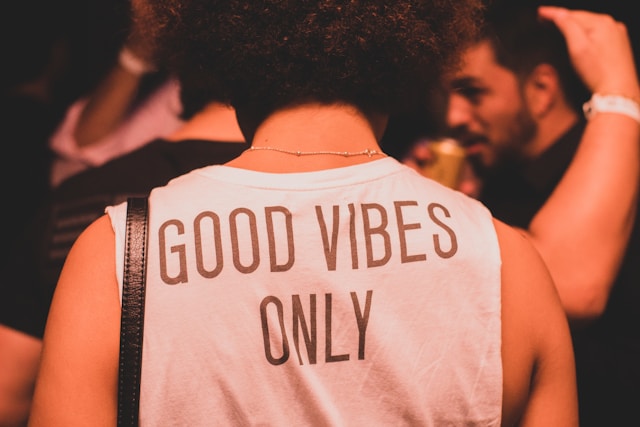 A person at a lively community gathering wearing a "Good Vibes Only" shirt, captured in warm, amber lighting to illustrate a Post-Parade Friendsgiving atmosphere.