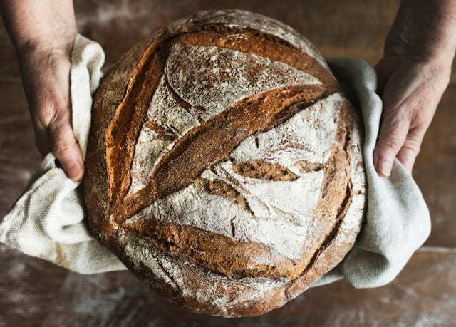 A top-down, close-up view of a large, crusty loaf of rustic sourdough bread being held by two hands in a white linen cloth. The golden-brown crust features artisanal flour-dusted scores, set against a warm, dark wooden background.