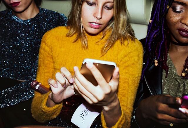 Three girls in a car and one blond woman is texting on her phone holding a bottle of wine.