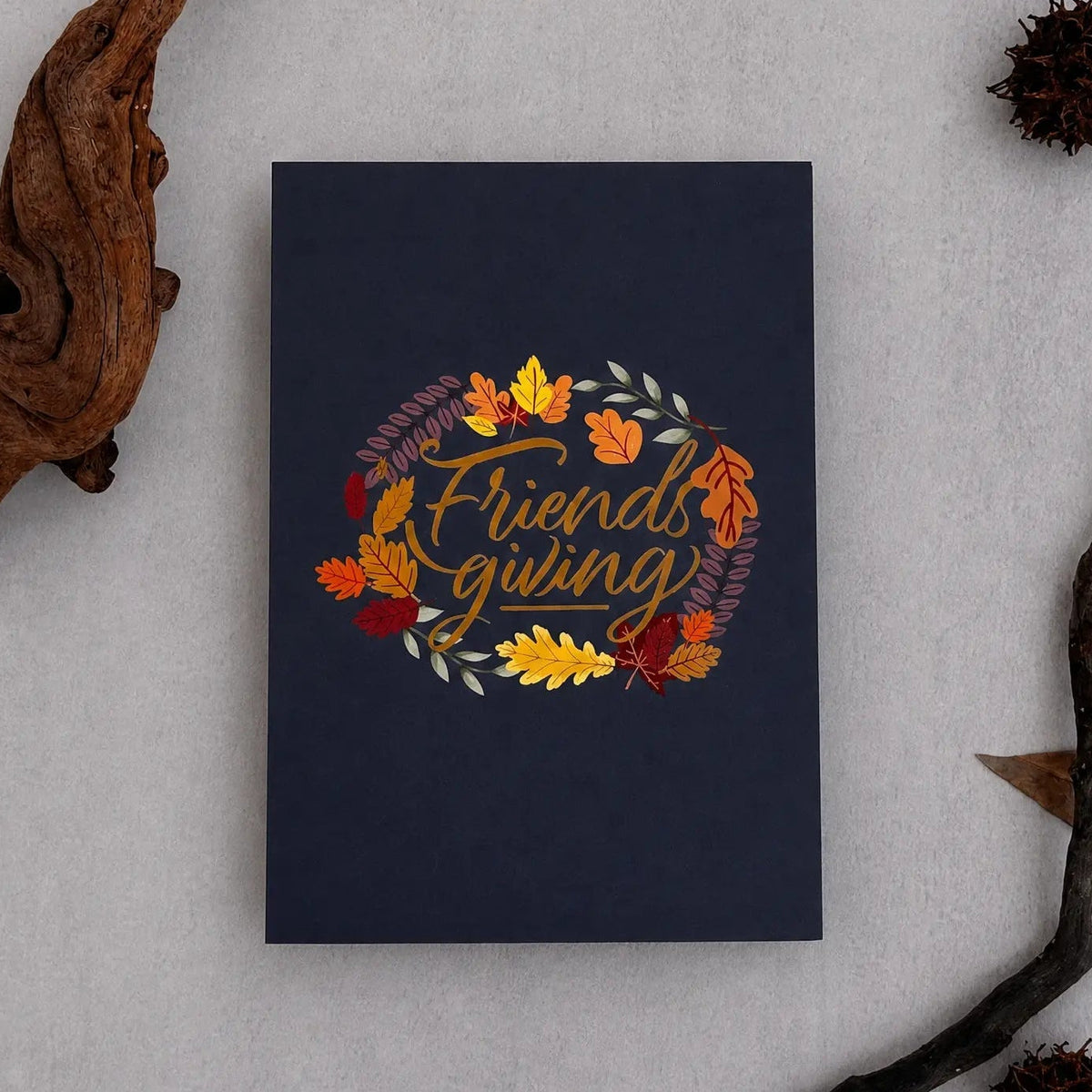 A navy blue greeting card with a 'Friends giving' autumn wreath design, styled in a flat-lay with driftwood and pinecones on a light gray textured background.