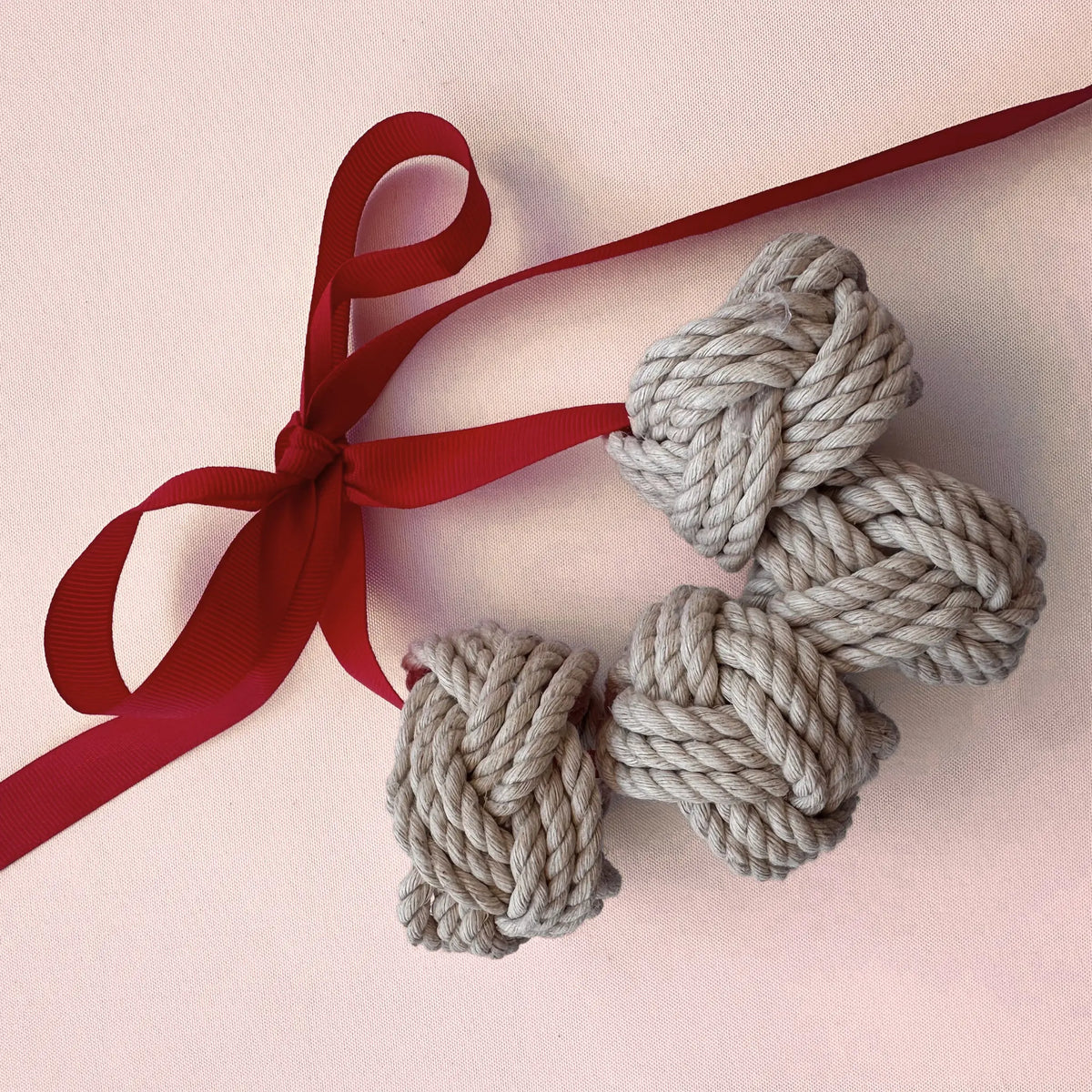 Three knotted gray rope ornaments tied to a red ribbon, arranged on a light pink background.