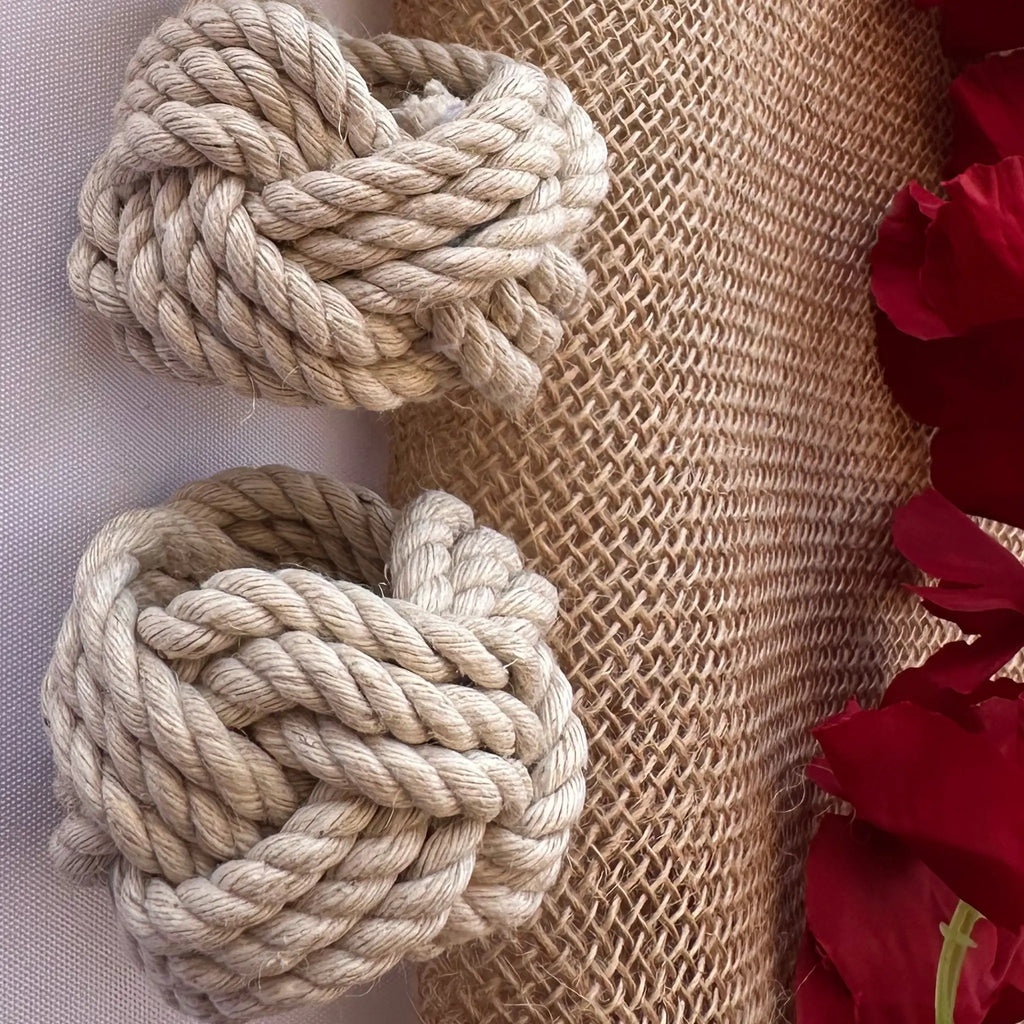 Two beige rope knotted loops arranged on a burlap and fabric backdrop with red rose petals along the right edge.