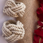 Two beige rope knotted loops arranged on a burlap and fabric backdrop with red rose petals along the right edge.