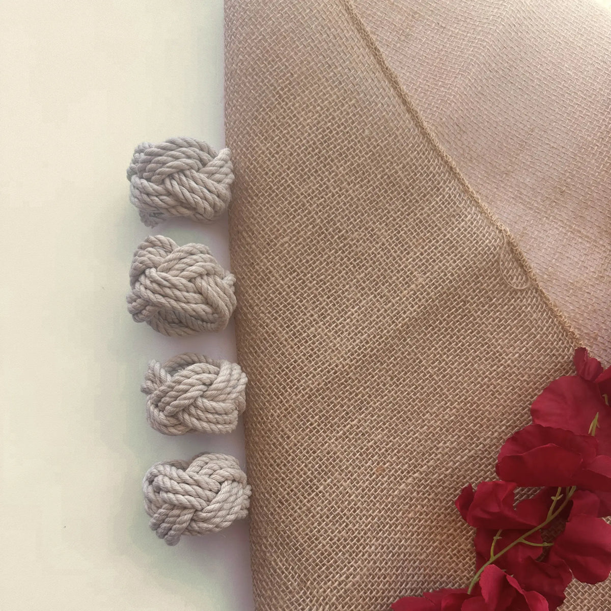 Four knotted gray rope tassels arranged vertically on the left side of a beige fabric panel, next to a white background and red flowers in the bottom right corner.