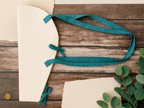 Teal ribbon tied around a white envelope on a wooden surface with green leaves.