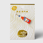 A 18K gold hairpin by Yellow Owl Workshop, shaped like a miniature red and blue bottle, presented on its illustrated white and gold backing card.