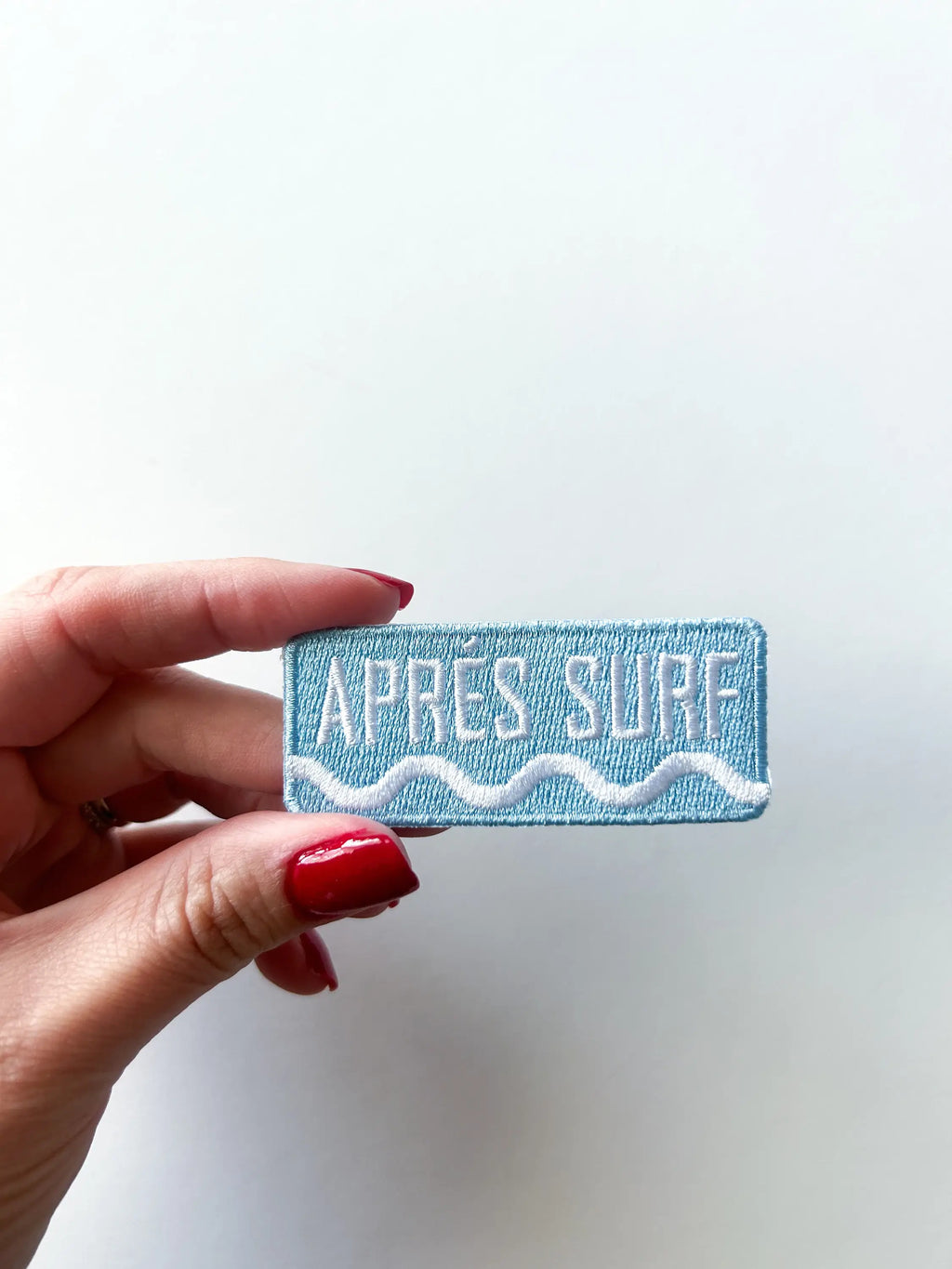 Hand holding a blue patch with 'APRES SURF' text against a white background