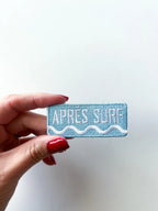 Hand holding a blue patch with 'APRES SURF' text against a white background
