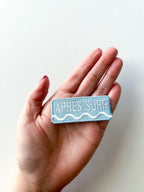 Hand holding a rectangular patch with 'APRèS SURF' text on a light background