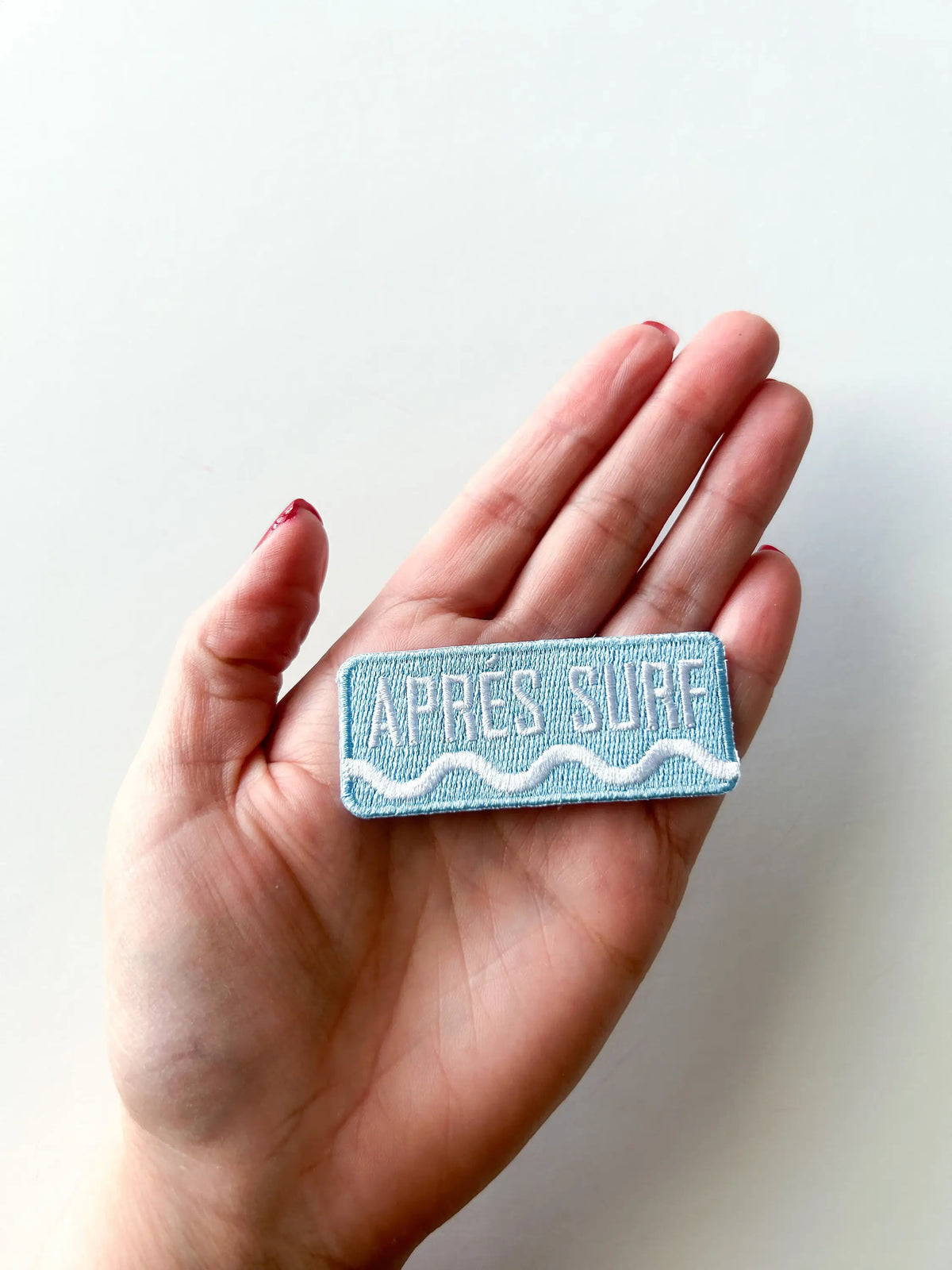 Hand holding a rectangular patch with 'APRèS SURF' text on a light background