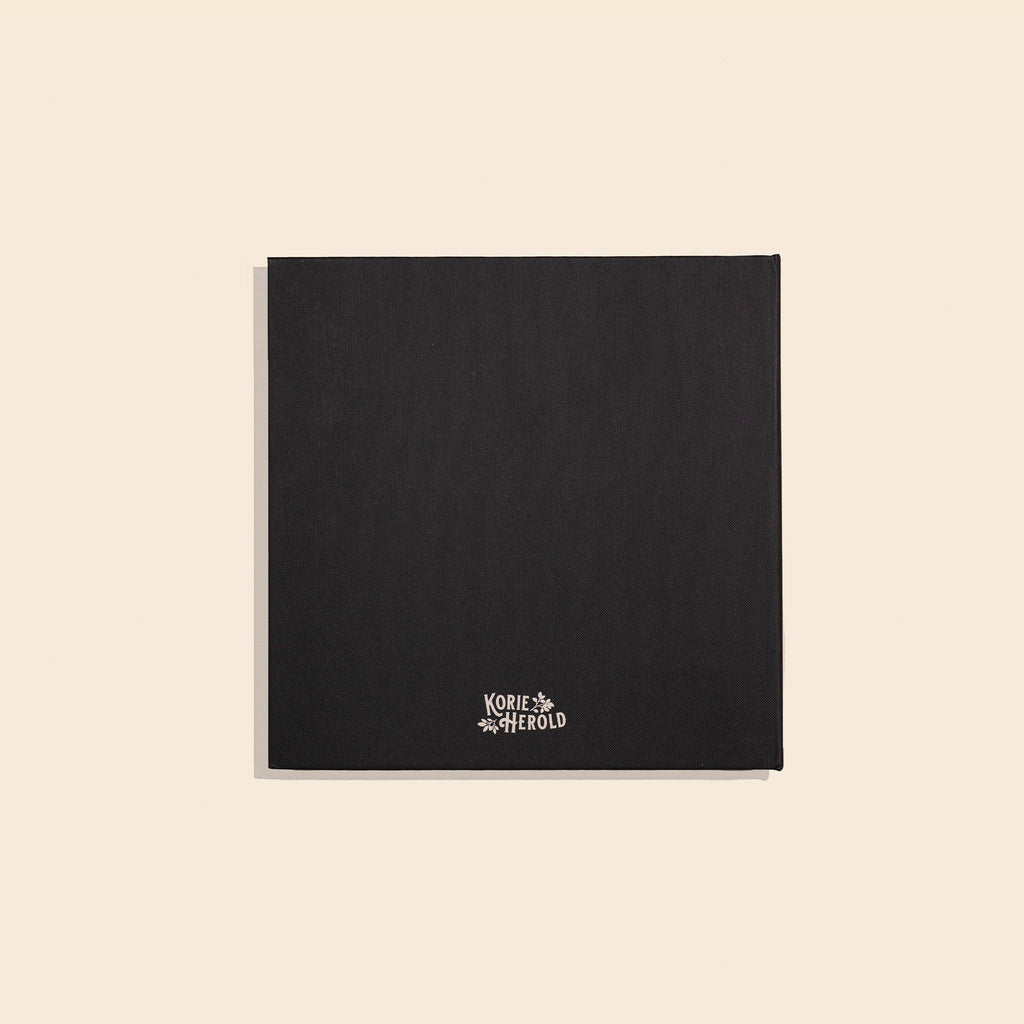 A black textured square card or notebook cover with the Korie & Aherold brand name printed in white at the bottom center, displayed on a solid light beige background.