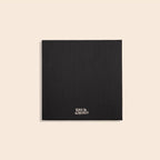 A black textured square card or notebook cover with the Korie & Aherold brand name printed in white at the bottom center, displayed on a solid light beige background.
