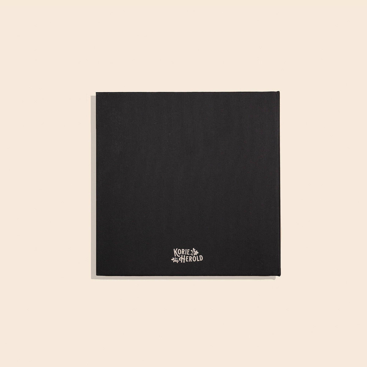 A black textured square card or notebook cover with the Korie & Aherold brand name printed in white at the bottom center, displayed on a solid light beige background.
