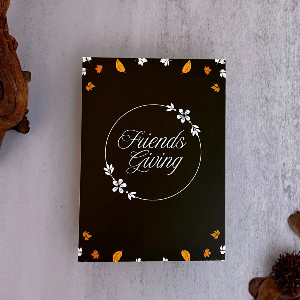 A black rectangular card or box with the words 'Friends Giving' in white script inside a circular floral border, styled on a light gray textured surface with dried leaves and driftwood.