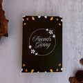 A black rectangular card or box with the words 'Friends Giving' in white script inside a circular floral border, styled on a light gray textured surface with dried leaves and driftwood.