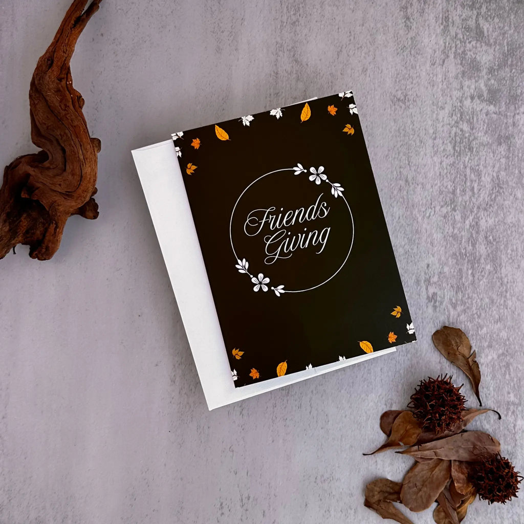 A square black greeting card with a white envelope, styled on a textured gray surface with dried brown leaves and branches. The card features the words 'Friends Giving' in white script inside a circular frame, decorated with small white flowers and orange and white leaf accents.