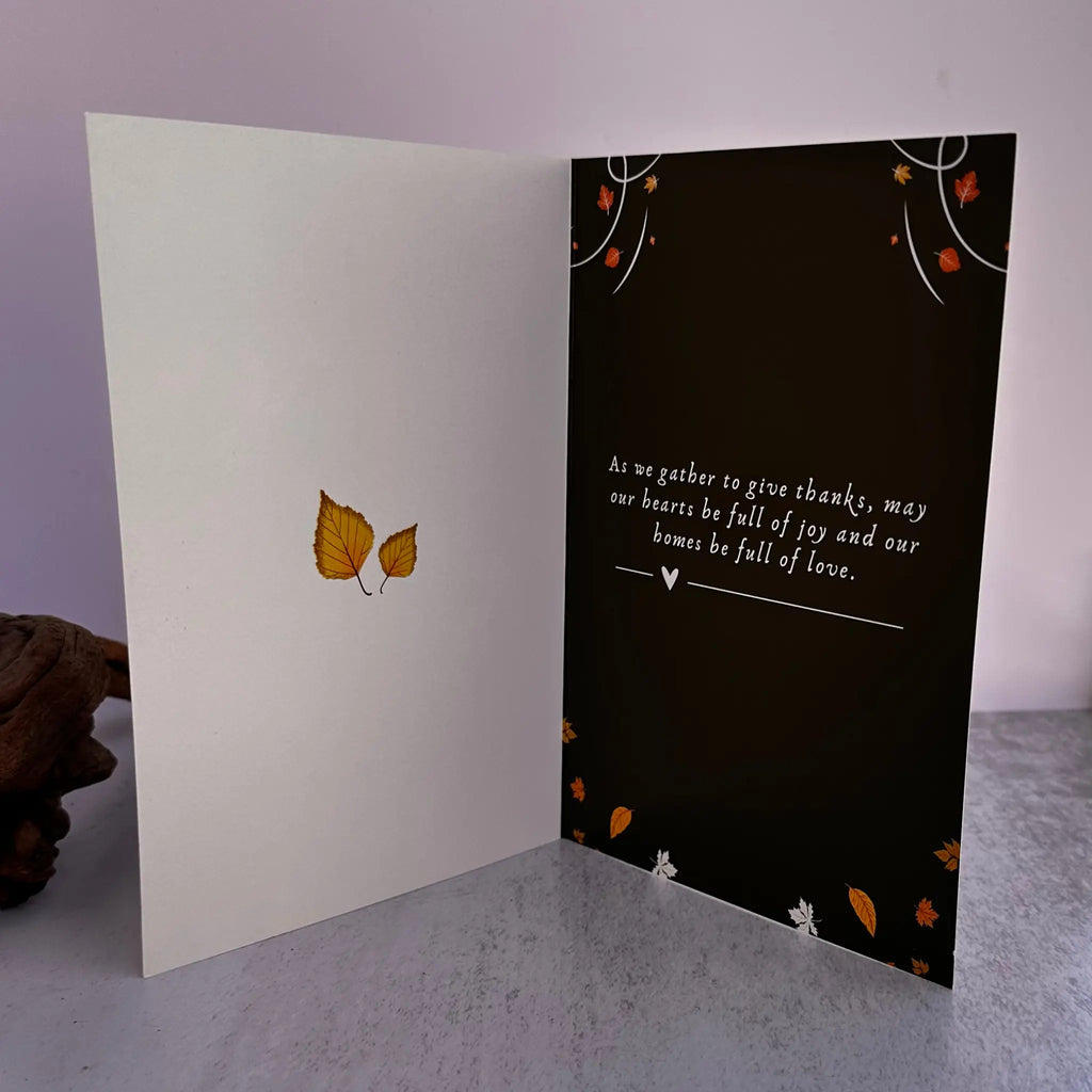 An open greeting card displayed on a gray surface against a pale lavender background. The left panel is white with two small yellow-orange leaves near the center, and the right panel is black with white script text and scattered orange and white leaf accents.