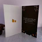 An open greeting card displayed on a gray surface against a pale lavender background. The left panel is white with two small yellow-orange leaves near the center, and the right panel is black with white script text and scattered orange and white leaf accents.
