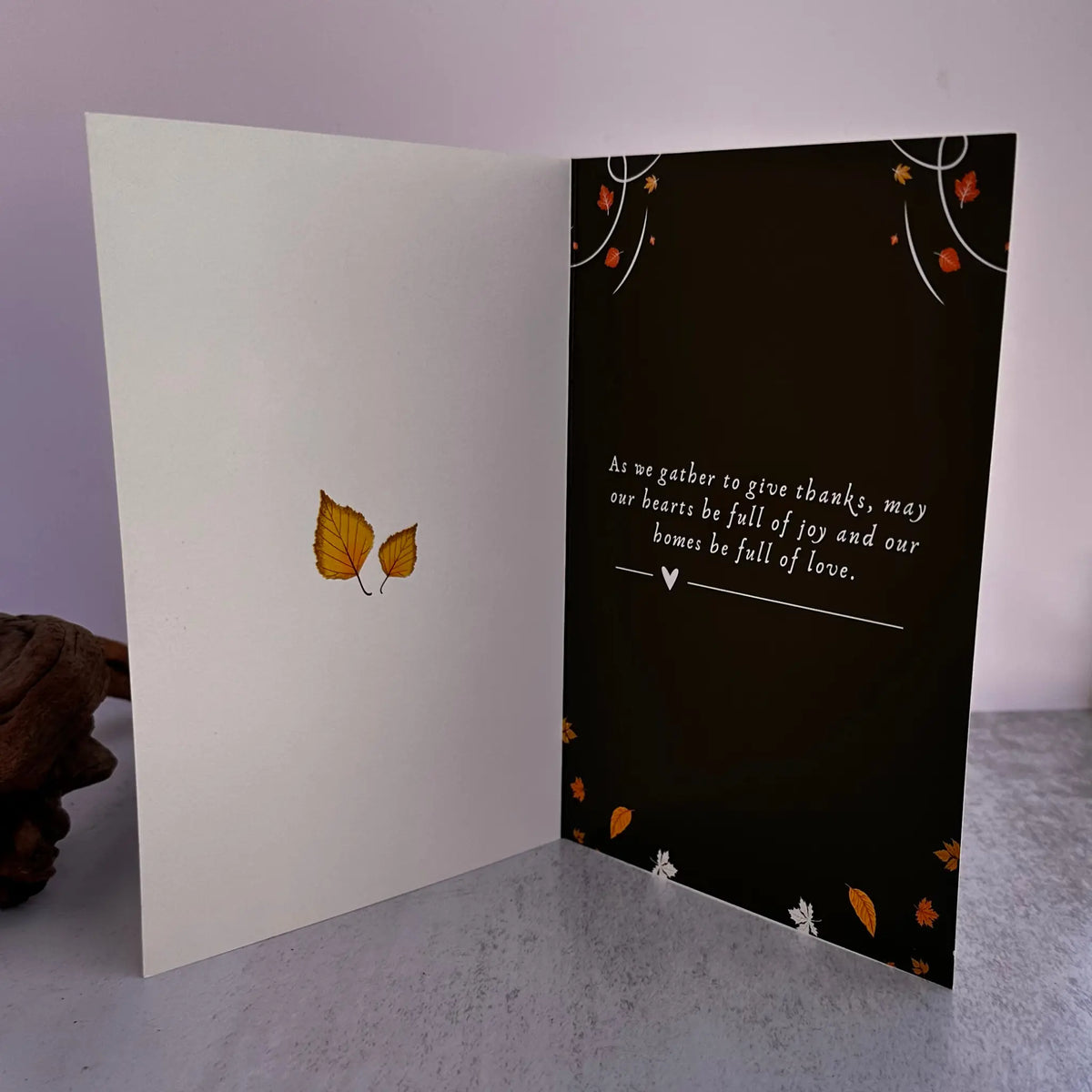 An open greeting card displayed on a gray surface against a pale lavender background. The left panel is white with two small yellow-orange leaves near the center, and the right panel is black with white script text and scattered orange and white leaf accents.