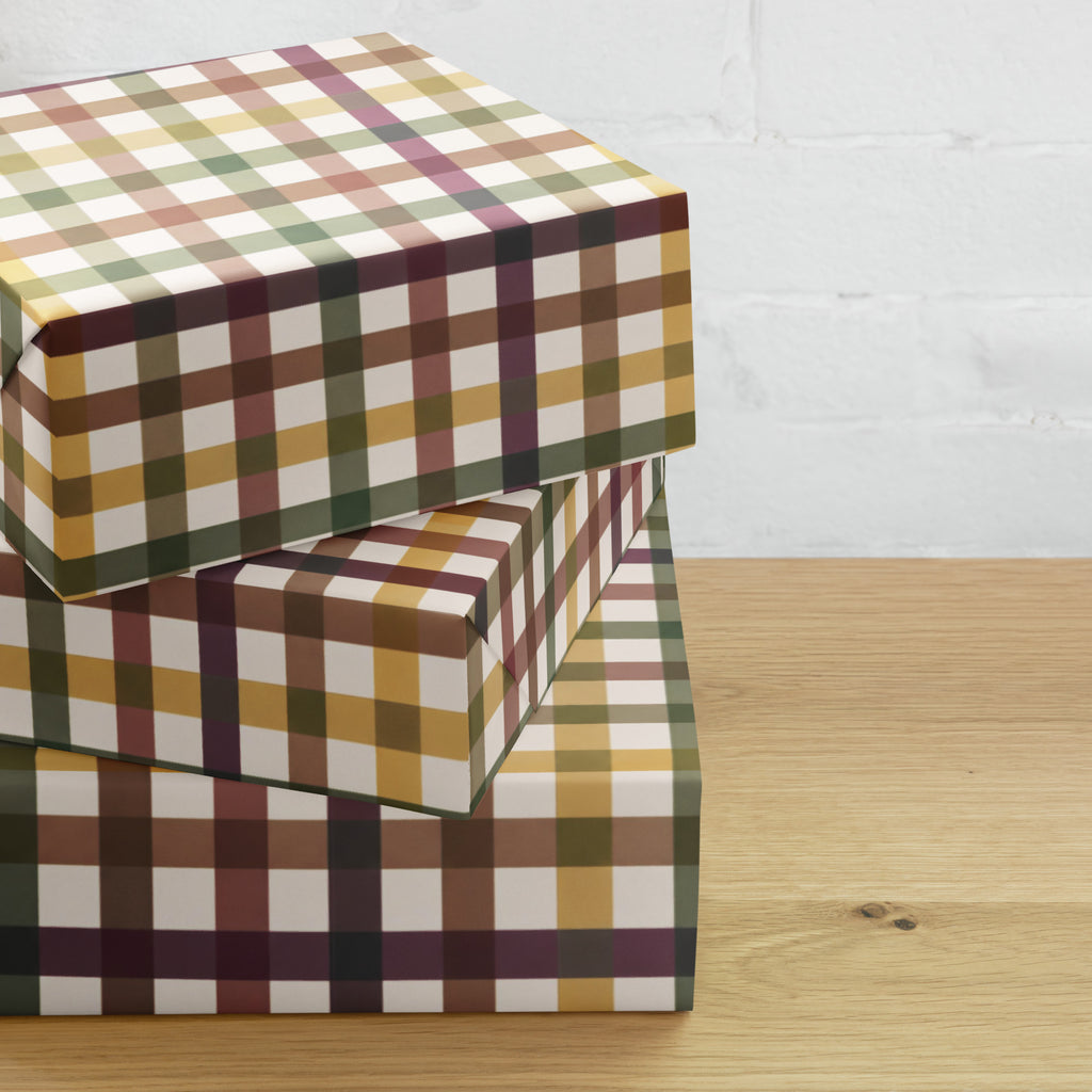 Three stacked square boxes wrapped in a plaid patterned paper, set on a light wood tabletop against a white brick wall background.