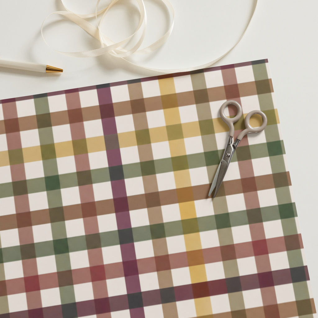 A styled flat-lay of a plaid wrapping paper sheet with a matching ribbon, a pair of scissors, and a pen on a clean white background.