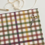 A styled flat-lay of a plaid wrapping paper sheet with a matching ribbon, a pair of scissors, and a pen on a clean white background.