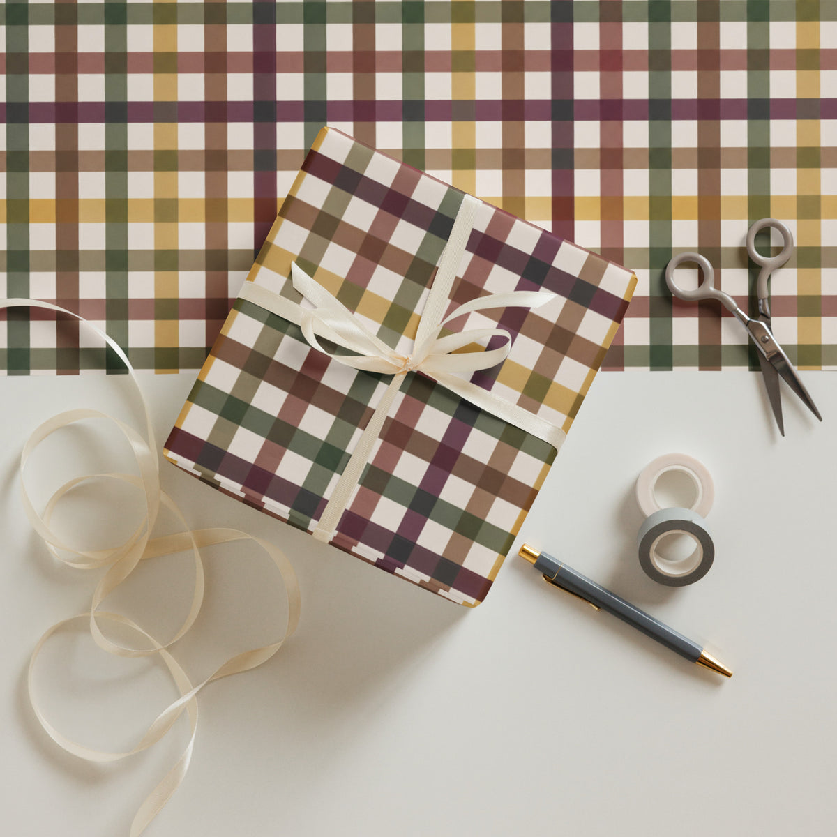 A wrapped gift box with a plaid patterned paper wrapping paper, tied with a cream ribbon, surrounded by gift-wrapping tools including scissors, tape rolls, and a pen on a light surface.