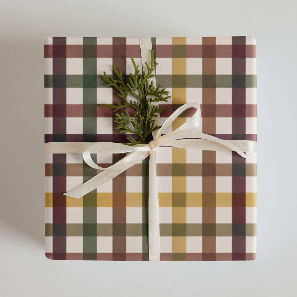 A square gift box wrapped in a plaid patterned paper with white, green, brown, and mustard yellow stripes, tied with a white satin ribbon and topped with a small green pine sprig, on a light gray background.