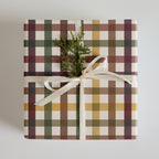 A square gift box wrapped in a plaid patterned paper with white, green, brown, and mustard yellow stripes, tied with a white satin ribbon and topped with a small green pine sprig, on a light gray background.