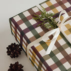 A wrapped gift box with a plaid pattern in olive green, mustard yellow, brown, and cream, tied with a white ribbon and decorated with a green pine sprig, with two pinecones on a light gray background.