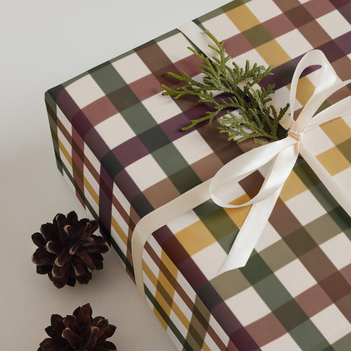 A wrapped gift box with a plaid pattern in olive green, mustard yellow, brown, and cream, tied with a white ribbon and decorated with a green pine sprig, with two pinecones on a light gray background.