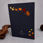 A dark navy blue greeting card featuring a decorative arrangement of colorful autumn leaves in orange, yellow, and red, displayed on a light gray surface with decorative driftwood.