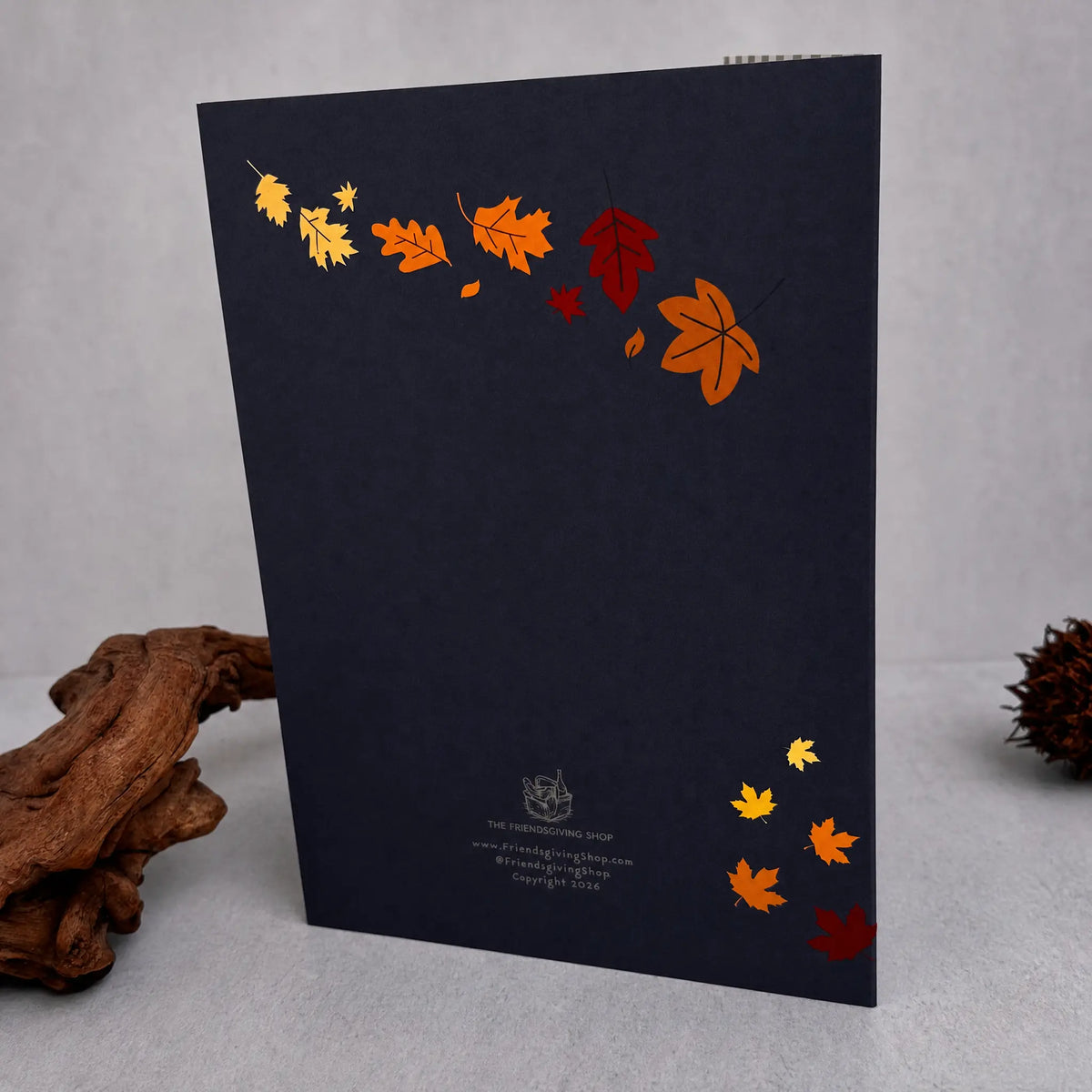 A dark navy blue greeting card featuring a decorative arrangement of colorful autumn leaves in orange, yellow, and red, displayed on a light gray surface with decorative driftwood.