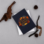 A navy blue greeting card with a circular autumn leaf wreath and the words 'Friends giving' in gold script, layered over a white card. The card is styled in a flat lay with dried branches, pinecones, and a seed pod on a light gray background.