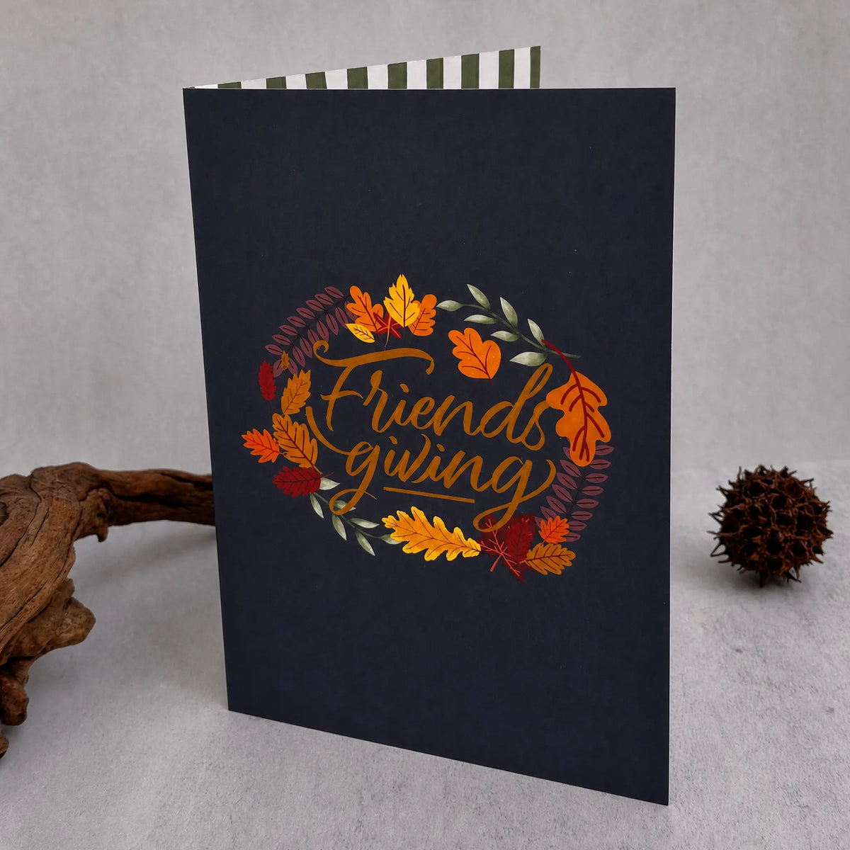 A dark navy blue greeting card standing upright on a light gray surface, decorated with an autumn wreath and the words 'Friends giving' in gold script. A small brown decorative branch and a pine cone sit beside the card, and part of a striped envelope is visible at the top.