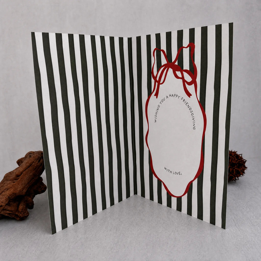 A folded greeting card with bold dark green and white vertical stripes, featuring a red outline ornament shape with a red ribbon bow and a blank white center for writing. The card is displayed open on a light gray surface with decorative driftwood props on either side.