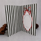 A folded greeting card with bold dark green and white vertical stripes, featuring a red outline ornament shape with a red ribbon bow and a blank white center for writing. The card is displayed open on a light gray surface with decorative driftwood props on either side.