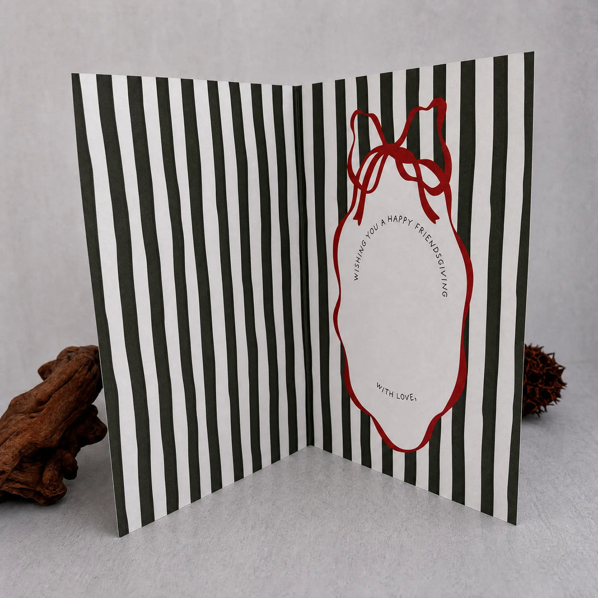 A folded greeting card with bold dark green and white vertical stripes, featuring a red outline ornament shape with a red ribbon bow and a blank white center for writing. The card is displayed open on a light gray surface with decorative driftwood props on either side.