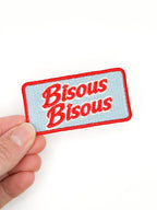 Embroidered patch with 'Bisous Bisous' text held by a hand on a white background