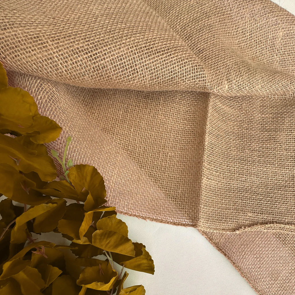 Natural tan burlap fabric with a coarse woven texture, draped to show its pattern and folds, with a decorative olive green leafy branch in the lower left corner.