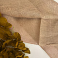 Natural tan burlap fabric with a coarse woven texture, draped to show its pattern and folds, with a decorative olive green leafy branch in the lower left corner.