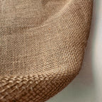 A close-up of woven natural burlap fabric showing its coarse, tan texture and folded edges against a light background.