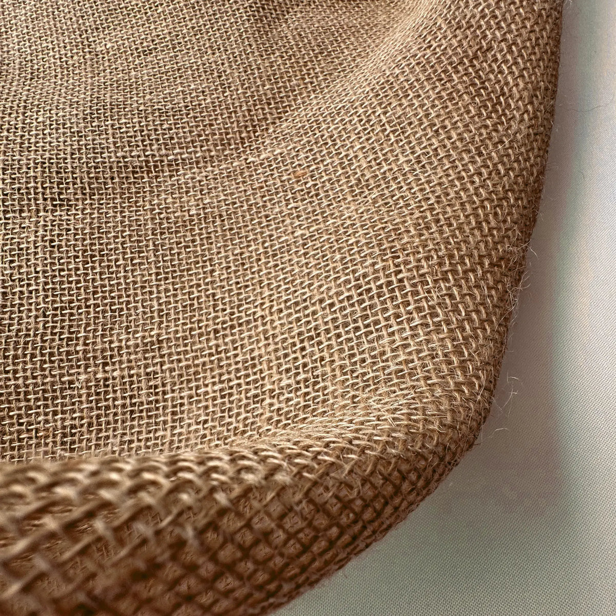 A close-up of woven natural burlap fabric showing its coarse, tan texture and folded edges against a light background.