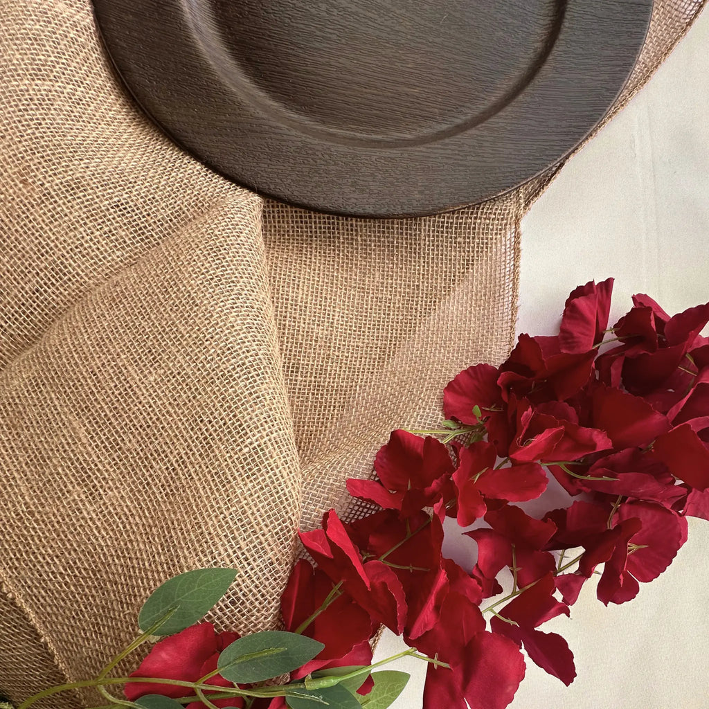 A close-up of a natural burlap table runner draped over a surface, styled with a dark brown wooden plate and red rose stems.