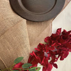 A close-up of a natural burlap table runner draped over a surface, styled with a dark brown wooden plate and red rose stems.