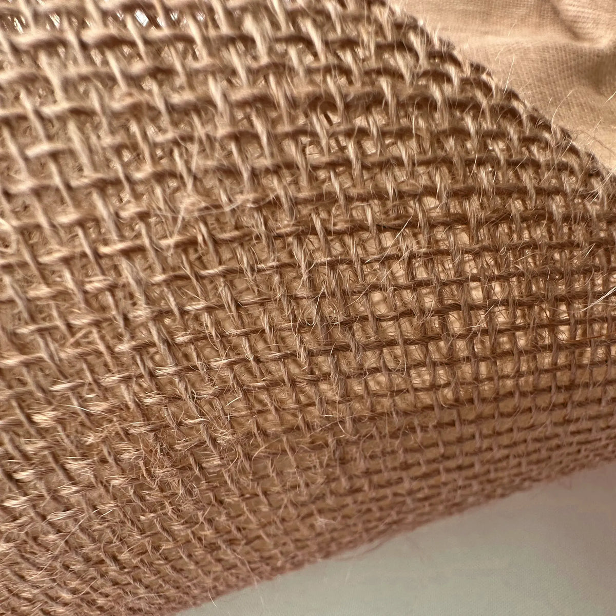 A close-up of a rolled piece of natural burlap fabric, showing its coarse woven texture and tan color, with a plain white surface visible underneath.