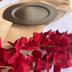 A dark gray ceramic plate resting on a beige burlap runner, with scattered deep red rose petals in the foreground on a white surface.